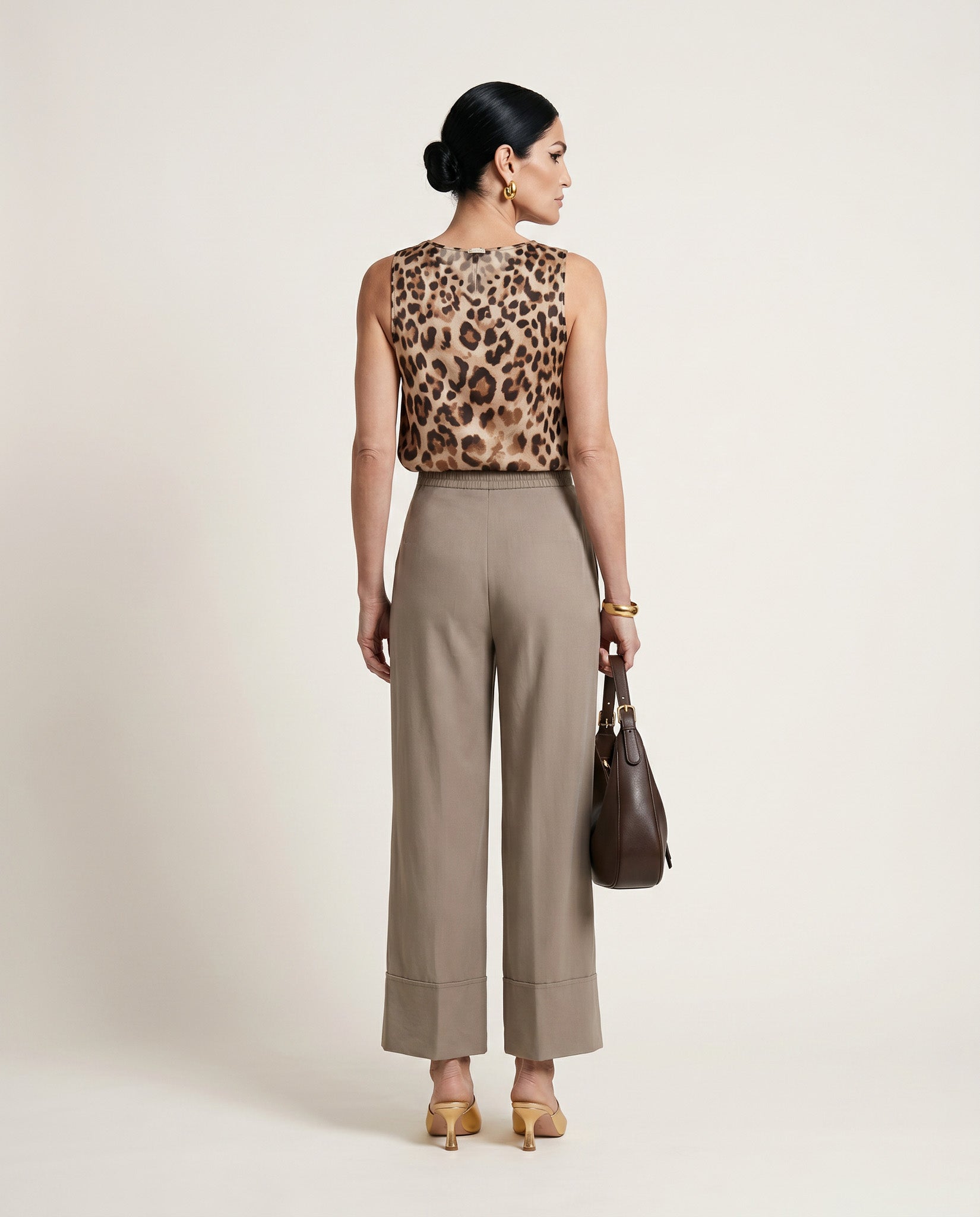 Woman wearing a leopard print top and beige pants with a brown handbag on a light background