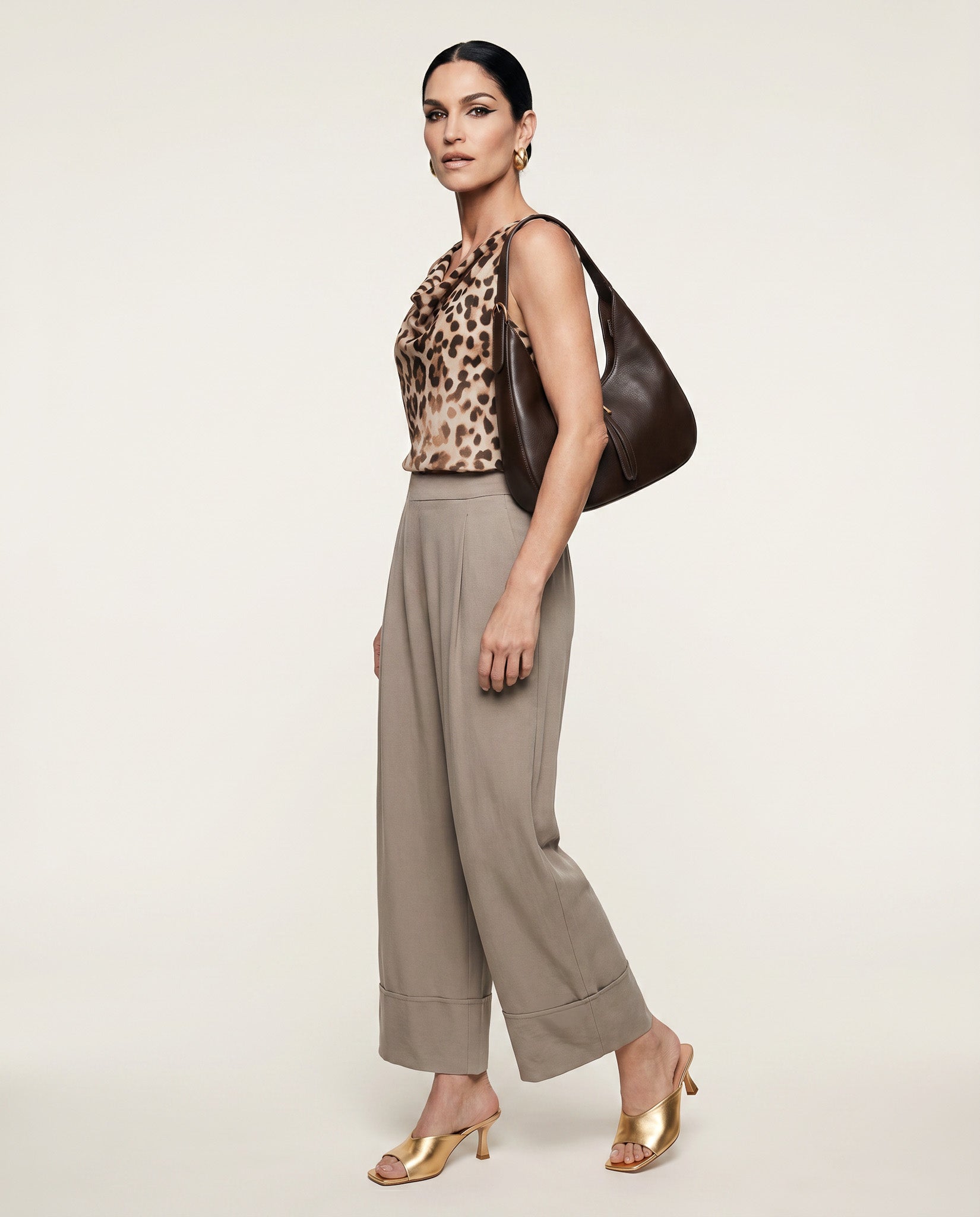 Woman wearing a leopard print top and beige pants, holding a brown handbag on a white background