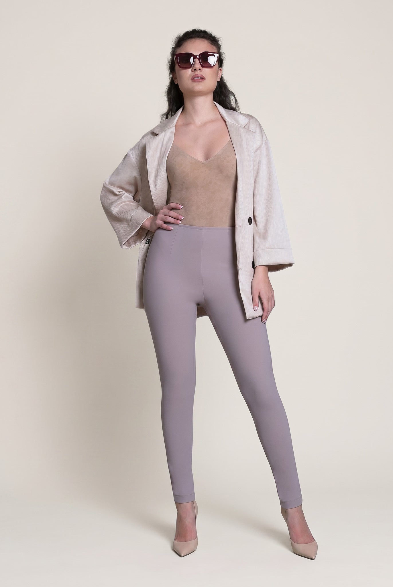 Woman wearing a beige top, light purple leggings, and a white jacket on a plain background