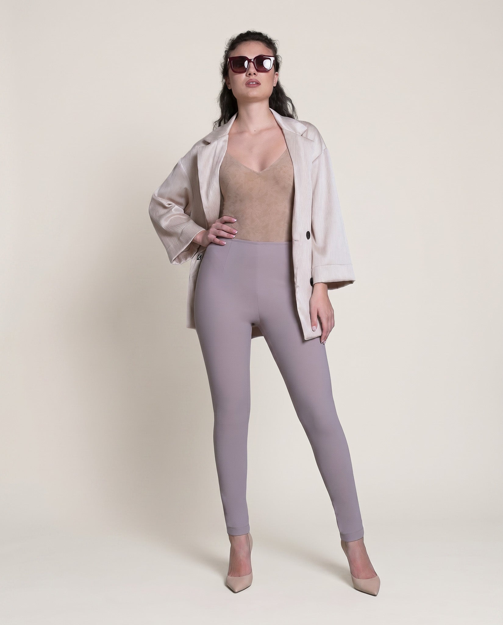 Woman wearing a beige top, light purple leggings, and a white jacket on a plain background